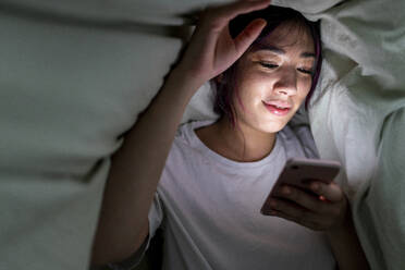 Smiling woman using smart phone under blanket at home - WPEF06843