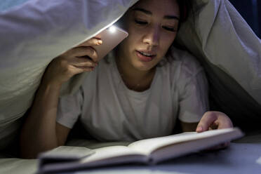 Young woman with smart phone reading book under blanket on bed at home - WPEF06844