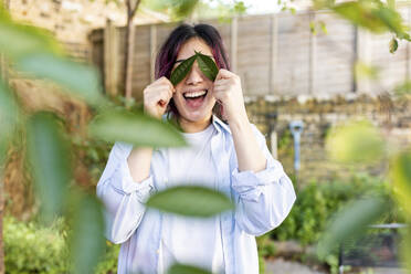 Fröhliche junge Frau hält Blätter über die Augen im Garten - WPEF06851