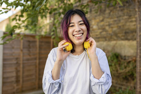 Glückliche junge Frau mit Kopfhörern im Garten stehend - WPEF06853