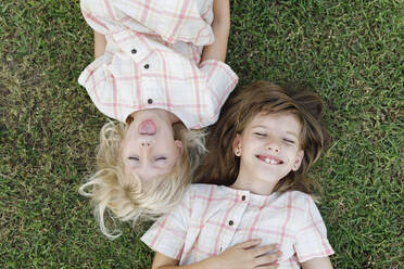 Cute girl sticking out tongue lying by happy sister on grass - TYF00486