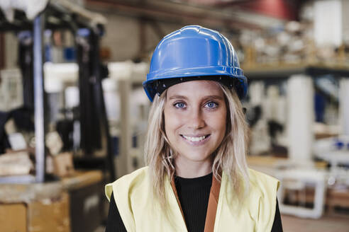Happy young engineer wearing hardhat standing in industry - EBBF07381