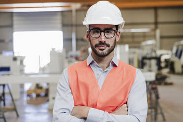 Selbstbewusster Ingenieur mit Brille und Arbeitsschutzkleidung, der mit verschränkten Armen in der Industrie steht - EBBF07402