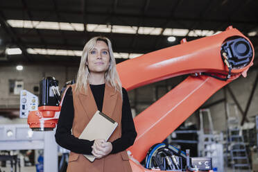 Lächelnder junger Ingenieur mit Tablet-PC vor einem Roboterarm stehend - EBBF07425