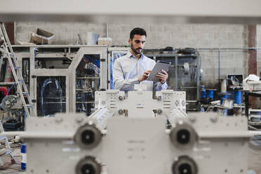 Businessman using tablet PC standing behind machine at industry - EBBF07439