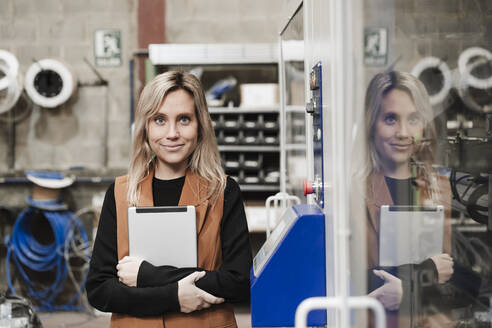 Smiling businesswoman with tablet PC standing by machine at industry - EBBF07443