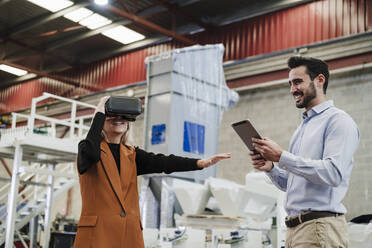 Glücklicher Geschäftsmann mit einem Kollegen, der einen Virtual-Reality-Simulator in einer Fabrik trägt - EBBF07447