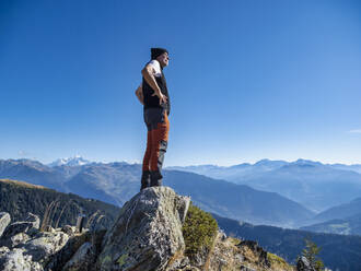 Aktiver älterer Mann mit Händen auf der Hüfte auf einem Felsen stehend unter blauem Himmel im Vanoise-Nationalpark, Frankreich - LAF02790