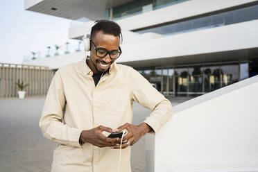 Happy man using smart phone wearing headphones outside building - RCPF01557