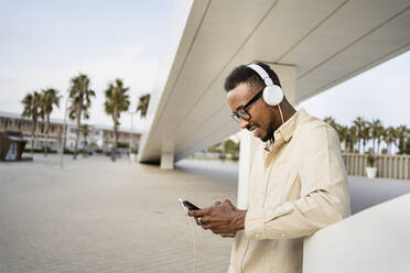 Man using mobile phone wearing headphones listening to music leaning by wall - RCPF01558