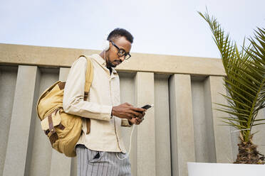 Man using smart phone with headphones standing by plant in front of wall - RCPF01565