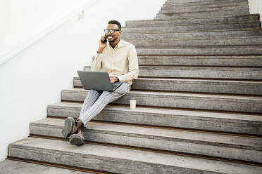 Smiling freelancer talking on smart phone sitting with laptop on steps - RCPF01571