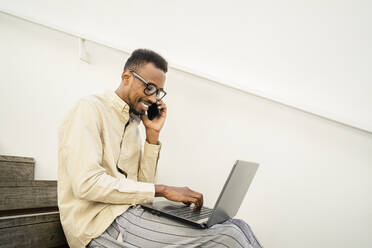 Smiling man talking on smart phone and using laptop by wall at stairs - RCPF01572