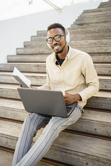 Glücklicher Mann sitzt mit Laptop und Kopfhörern auf einer Treppe - RCPF01573