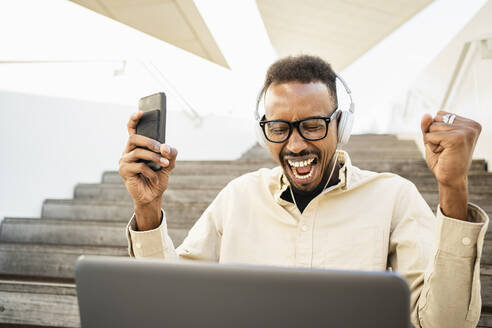 Excited freelancer with laptop and smart phone sitting on steps - RCPF01576