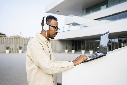 Freelancer wearing headphones using laptop outside building - RCPF01587