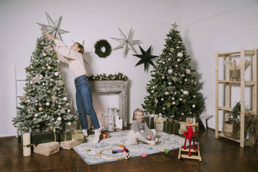 Mutter schmückt Weihnachtsbaum mit niedlichen Tochter sitzt auf Teppich zu Hause - NDEF00244