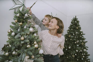 Lächelnde Mutter, die ihre Tochter huckepack nimmt, um einen Stern am Weihnachtsbaum anzubringen - NDEF00257