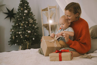 Mother helping son unwrapping Christmas gift on bed at home - NDEF00274