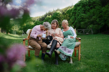 Happy senior women friends sitting on a bench and drinking tea outdoors in garden, laughing. - HPIF03148