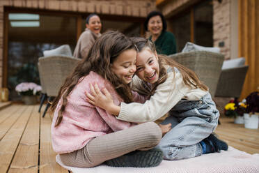 Two happy little sisters hugging outdoors in a patio in autumn, mother and grandmother at background. - HPIF03168