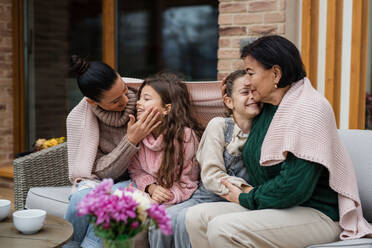 Zwei glückliche Schwestern mit einer Mutter und einer Großmutter sitzen eingewickelt in eine Decke im Freien im Innenhof im Herbst - HPIF03171