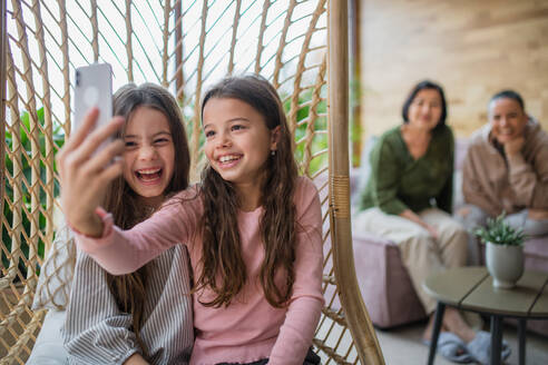 Glückliche kleine Schwestern sitzen in einem Rattan-Hängesessel im Wintergarten und machen ein Selfie. - HPIF03186