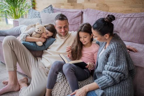 Zwei glückliche Schwestern mit Mutter und Vater sitzen auf dem Boden, umarmen sich und lesen ein Buch zu Hause - HPIF03197