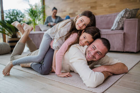 Two happy sisters having fun with a father, lying on his back with mother at background at home - HPIF03211