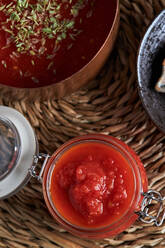 Draufsicht auf eine Pfanne und einen Behälter mit köstlicher hausgemachter Tomatensoße, bestreut mit Oregano, auf einer gewebten Matte auf einem Holztisch in der Küche - ADSF41520