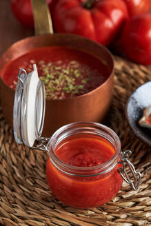 Draufsicht auf eine Pfanne und einen Behälter mit köstlicher hausgemachter Tomatensoße, bestreut mit Oregano, auf einer gewebten Matte auf einem Holztisch in der Küche - ADSF41521