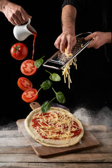 Koch reibt Käse und gießt Marinara-Sauce, während Tomaten und Basilikumblätter auf rohe Pizza vor schwarzem Hintergrund fallen - ADSF41545