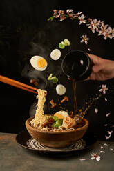 Cook holding noodles with chopsticks and adding miso broth into ramen while eggs and onion with mushrooms falling into bowl - ADSF41546