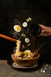 Cook holding noodles with chopsticks and adding miso broth into ramen while eggs and onion with mushrooms falling into bowl - ADSF41548