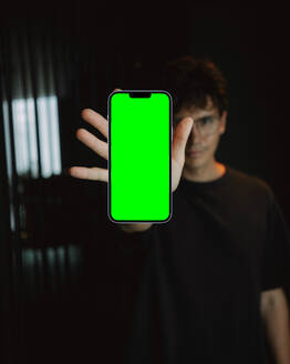 Weichzeichner des Mobiltelefons mit chromakey grünem Bildschirm in der Hand eines jungen lockigen Mannes mit Brille - ADSF41578