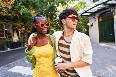 Cheerful confident young multiethnic couple in stylish outfits and sunglasses holding hands while crossing paved road on city street - ADSF41583