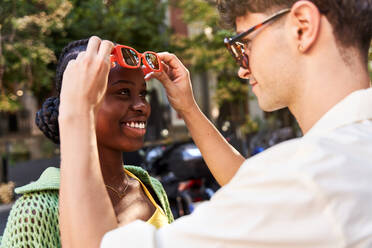 Seitenansicht von Ernte verliebten jungen hispanischen Mann in weißem Hemd lächelnd und Anpassung Sonnenbrille der fröhlichen afroamerikanischen Freundin auf der Straße der Stadt an einem sonnigen Tag - ADSF41585