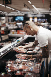 Seitenansicht eines tätowierten männlichen Kunden in Freizeitkleidung, der in der Nähe einer Gefriertruhe steht und ein Steak für das Abendessen im Supermarkt auswählt - ADSF41595