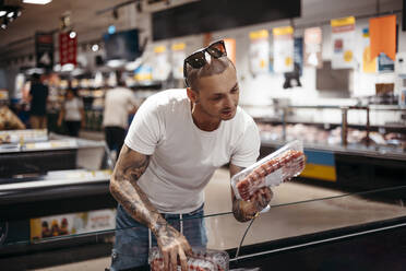 Tätowierter männlicher Kunde in Freizeitkleidung steht in der Nähe der Gefriertruhe und wählt ein Steak für das Abendessen im Supermarkt aus - ADSF41596