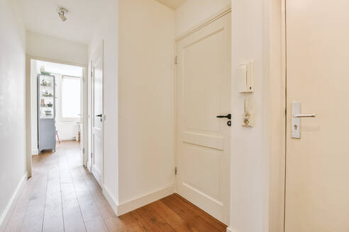 Schmaler Korridor mit weißen Wänden und Türen, die zu einem geräumigen Zimmer mit Fenstern und Parkettboden in einer modernen Wohnung führen - ADSF41607