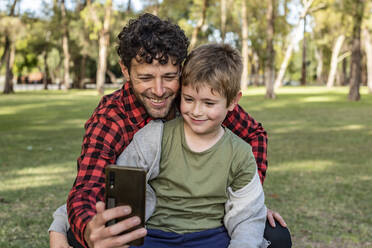 Positive man taking self portrait on smartphone together with glad preteen son while sitting on grass in forest in summer - ADSF41698