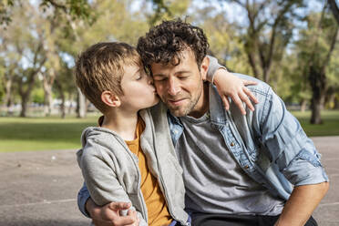 Son in casual clothes hugging and kissing dad on cheek while spending sunny weekend day in park - ADSF41701