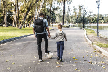 Rückenansicht eines anonymen Vaters und eines Sohnes mit weißem Ball, die sich an den Händen halten und auf einem Pfad in der Nähe eines Sees an einem Herbstwochenende im Park spazieren gehen - ADSF41702