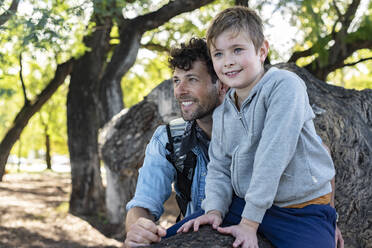 Happy son sitting on tree trunk near optimistic dad and looking away while spending weekend day in park together - ADSF41706