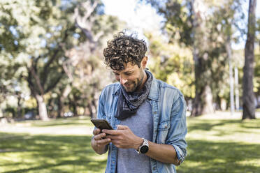 Mann in Freizeitkleidung, der an einem sommerlichen Wochenendtag in einem sonnenbeschienenen Park sein Mobiltelefon benutzt - ADSF41713