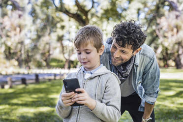 Glad dad smiling and watching son playing videogame on cellphone while spending sunny weekend day in park together - ADSF41714