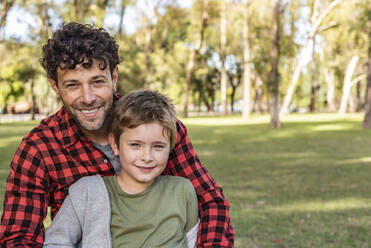 Happy father in checkered clothes smiling and hugging laughing son while sitting on grassy lawn on sunny weekend day in park - ADSF41715