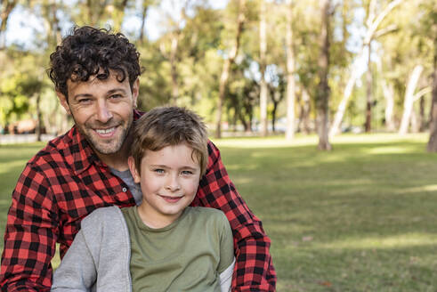 Glücklicher Vater in karierter Kleidung, der lächelnd seinen lachenden Sohn umarmt, während er an einem sonnigen Wochenendtag im Park auf einer Wiese sitzt - ADSF41715