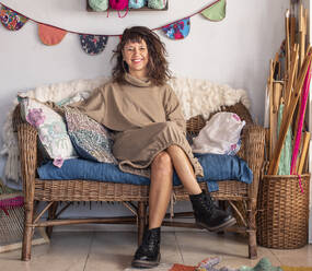 Full body of smiling adult female loom weaver. in beige dress and boots sitting on sofa with crossed legs near wooden tools - ADSF41721