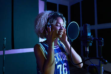 Side view of young blond woman in trendy outfit standing near microphone while recording song in studio with glowing purple lights - ADSF41723